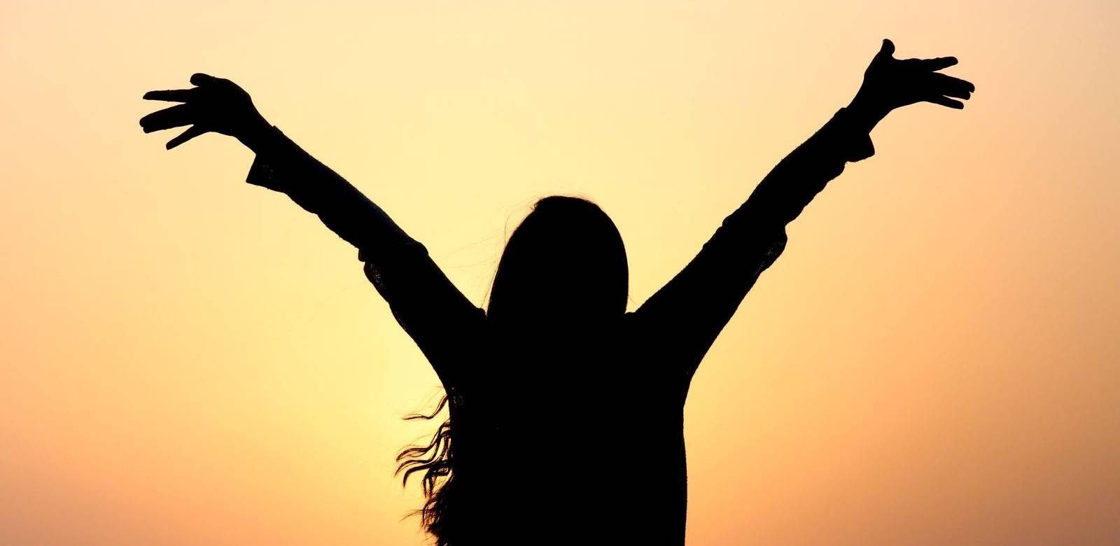 A woman standing with her arms raised toward a golden sunrise